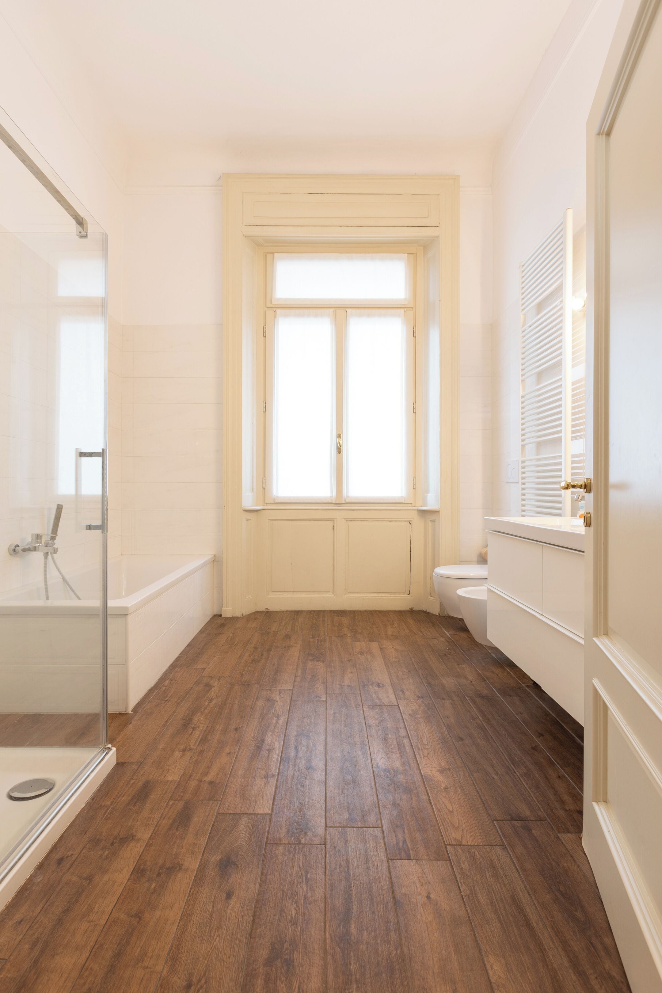 Une salle de bain avec un plancher en bois, une douche en verre, des toilettes blanches et une fenêtre avec des stores.