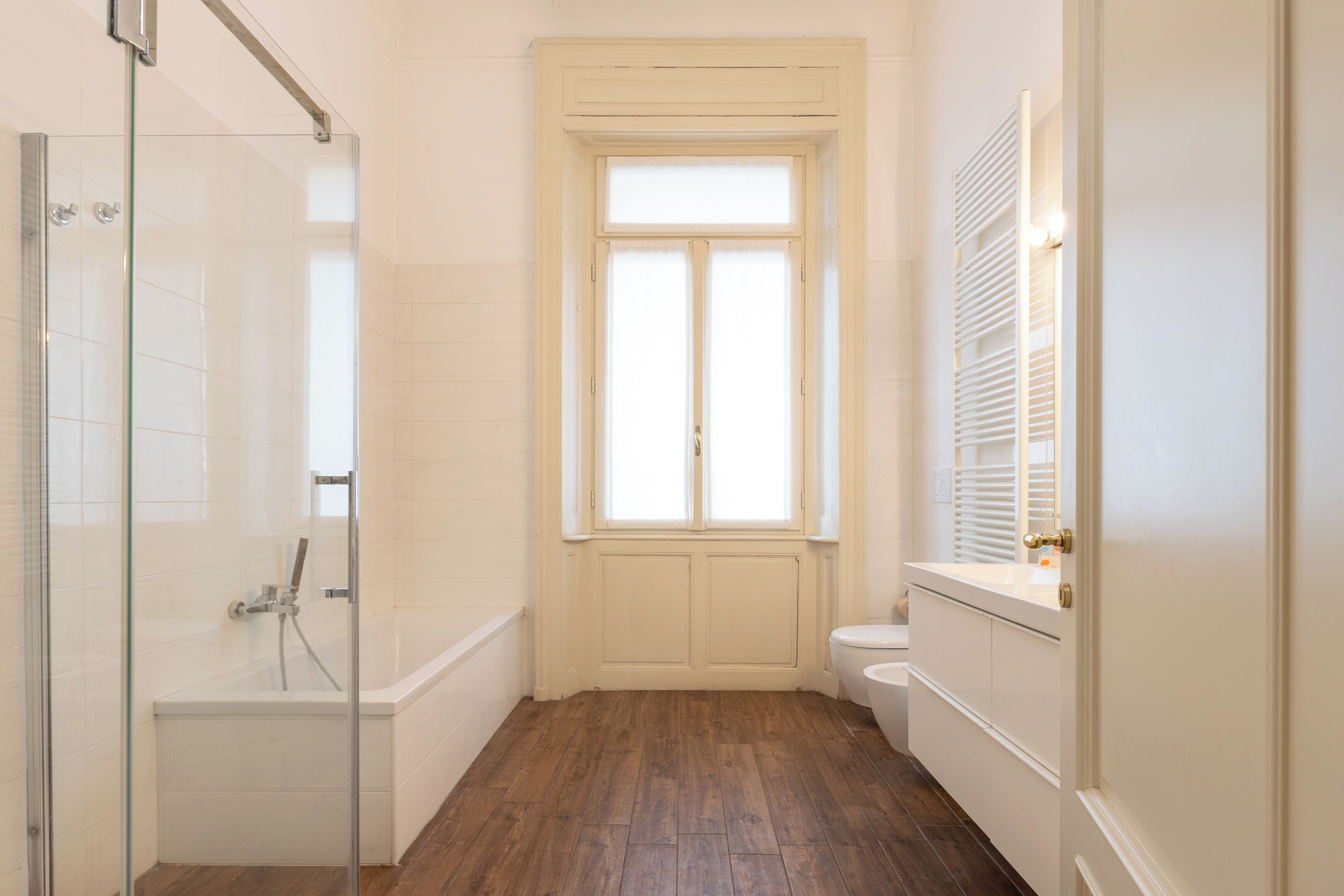 A bathroom with a glass shower enclosure, a wooden floor, a window with shutters, and a toilet.