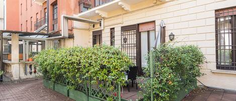 Un patio con un jardín verde, una zona de estar con sillas y un edificio con balcón.