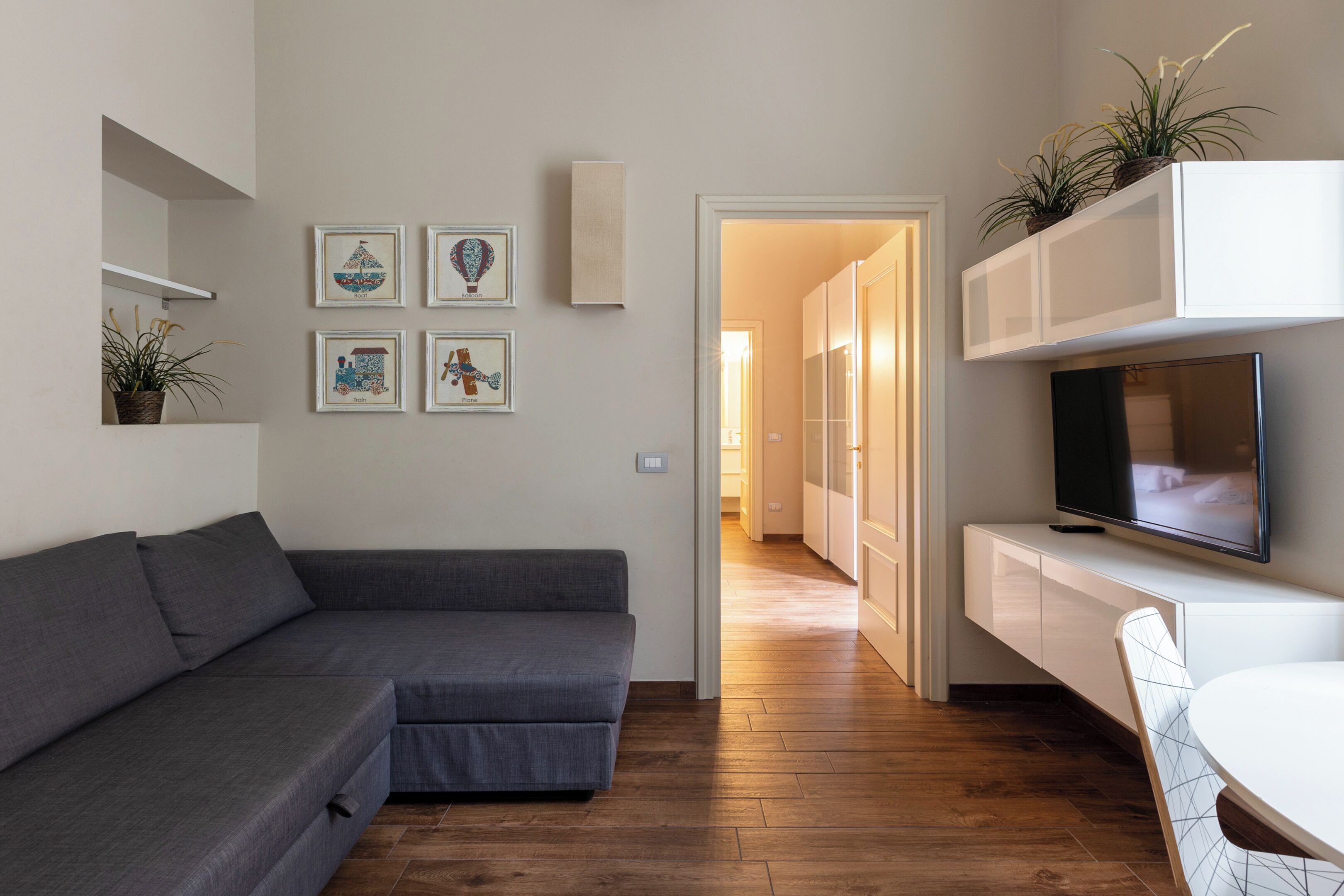 Uma sala moderna com um sofá seccional cinza, uma televisão de tela plana em um móvel branco, e uma área de jantar com mesa e cadeira brancas.