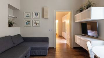 Uma sala moderna com um sofá seccional cinza, uma televisão de tela plana em um móvel branco, e uma área de jantar com mesa e cadeira brancas.