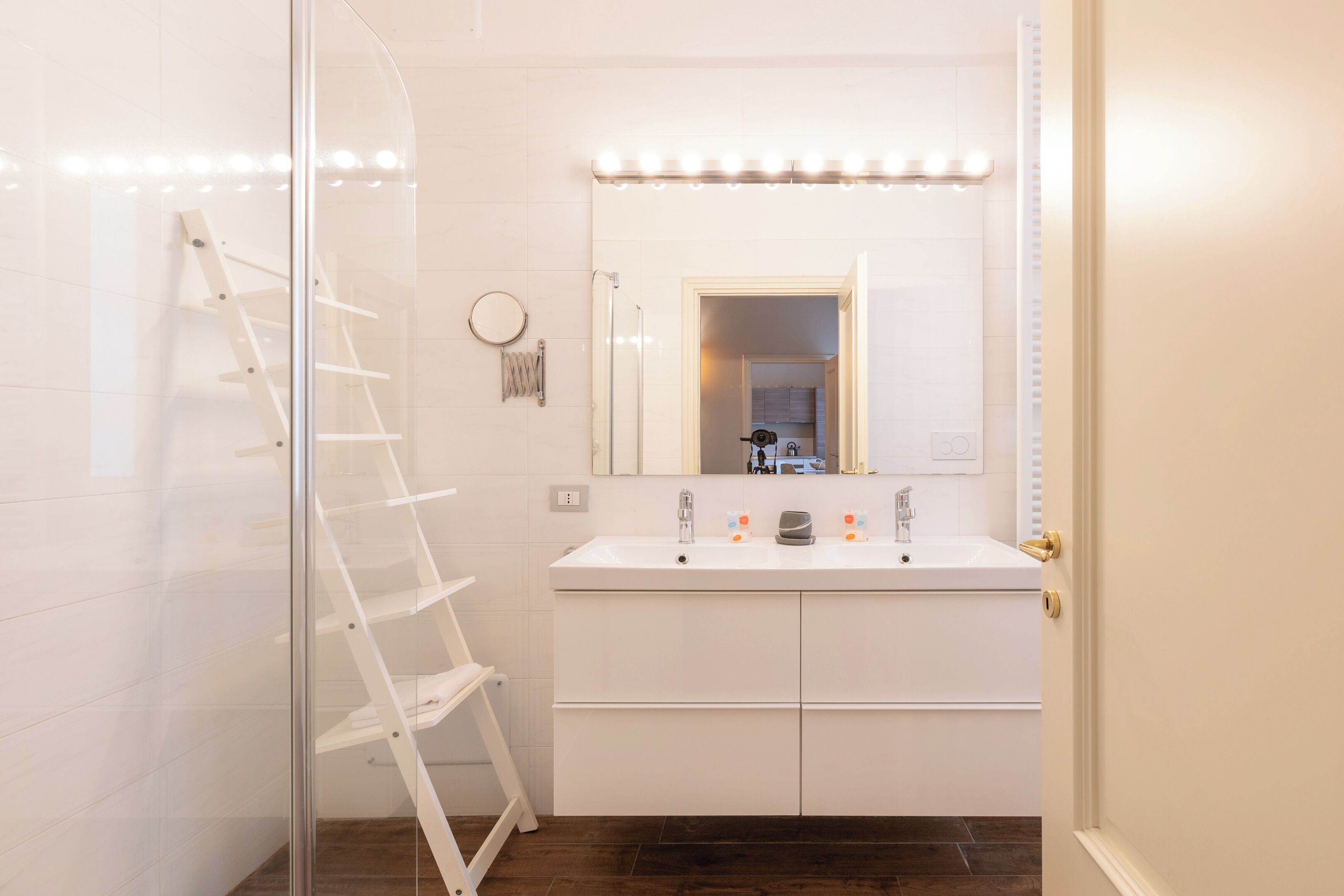 A bathroom with a white vanity, a mirror with lights, a ladder, and a shower area.