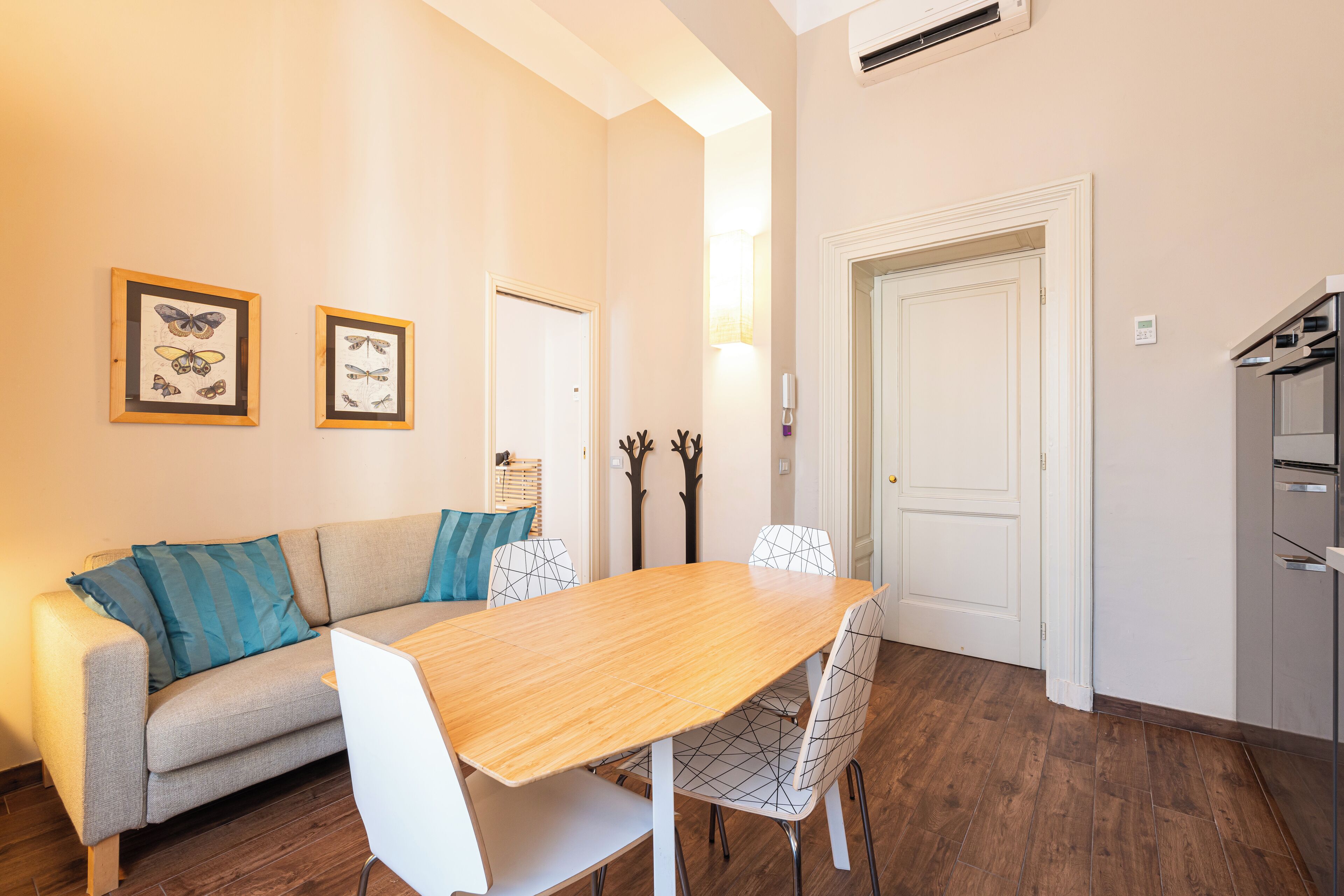 Un comedor con mesa de madera y sillas blancas, sofá gris con cojines azules y cuadros de mariposas en la pared.