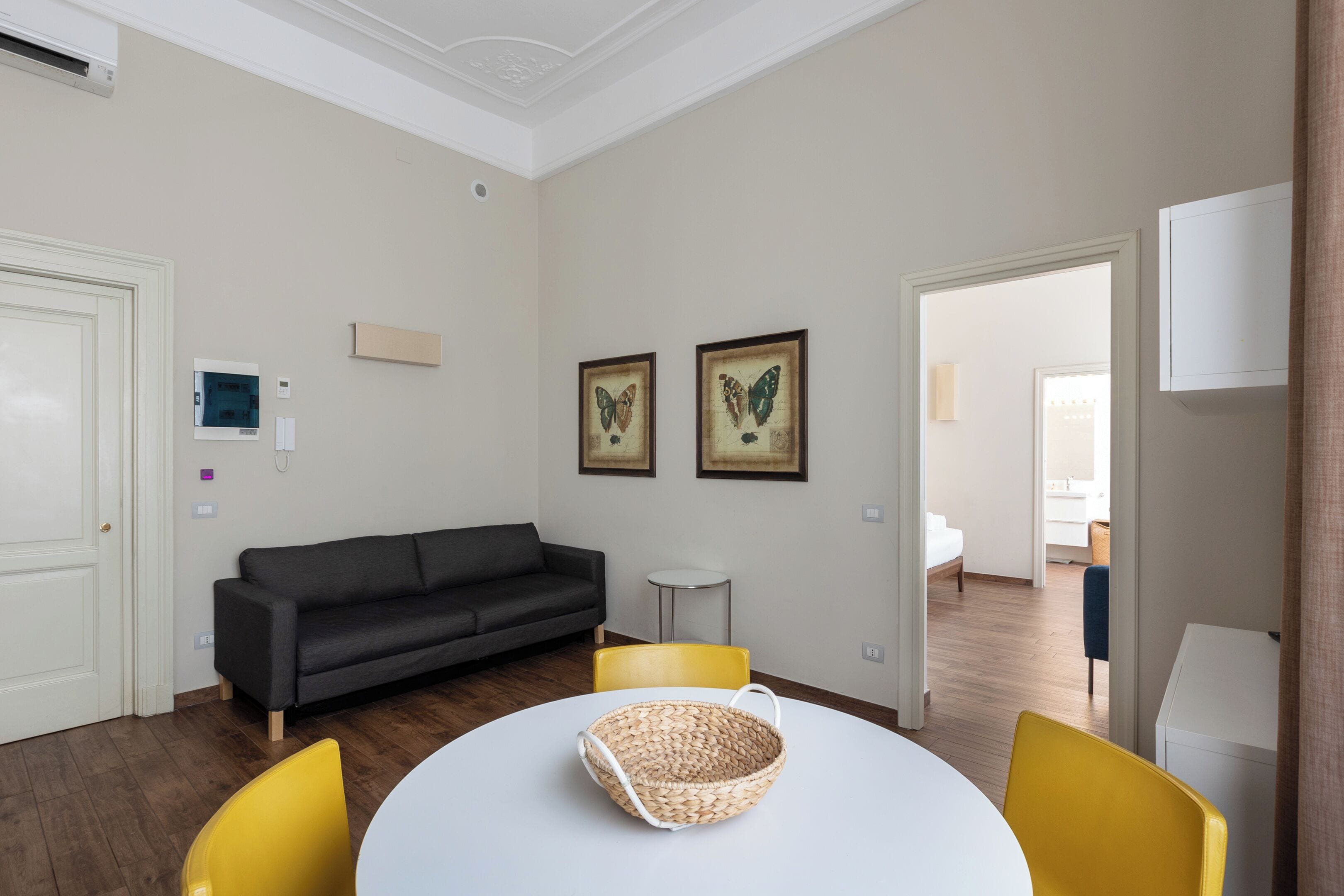 Un salon moderne avec un canapé gris foncé, une table ronde blanche surmontée d’un panier en osier et deux fauteuils jaunes. On peut voir deux tableaux encadrés représentant des papillons accrochés au mur, une petite table d’appoint blanche et un meuble blanc.