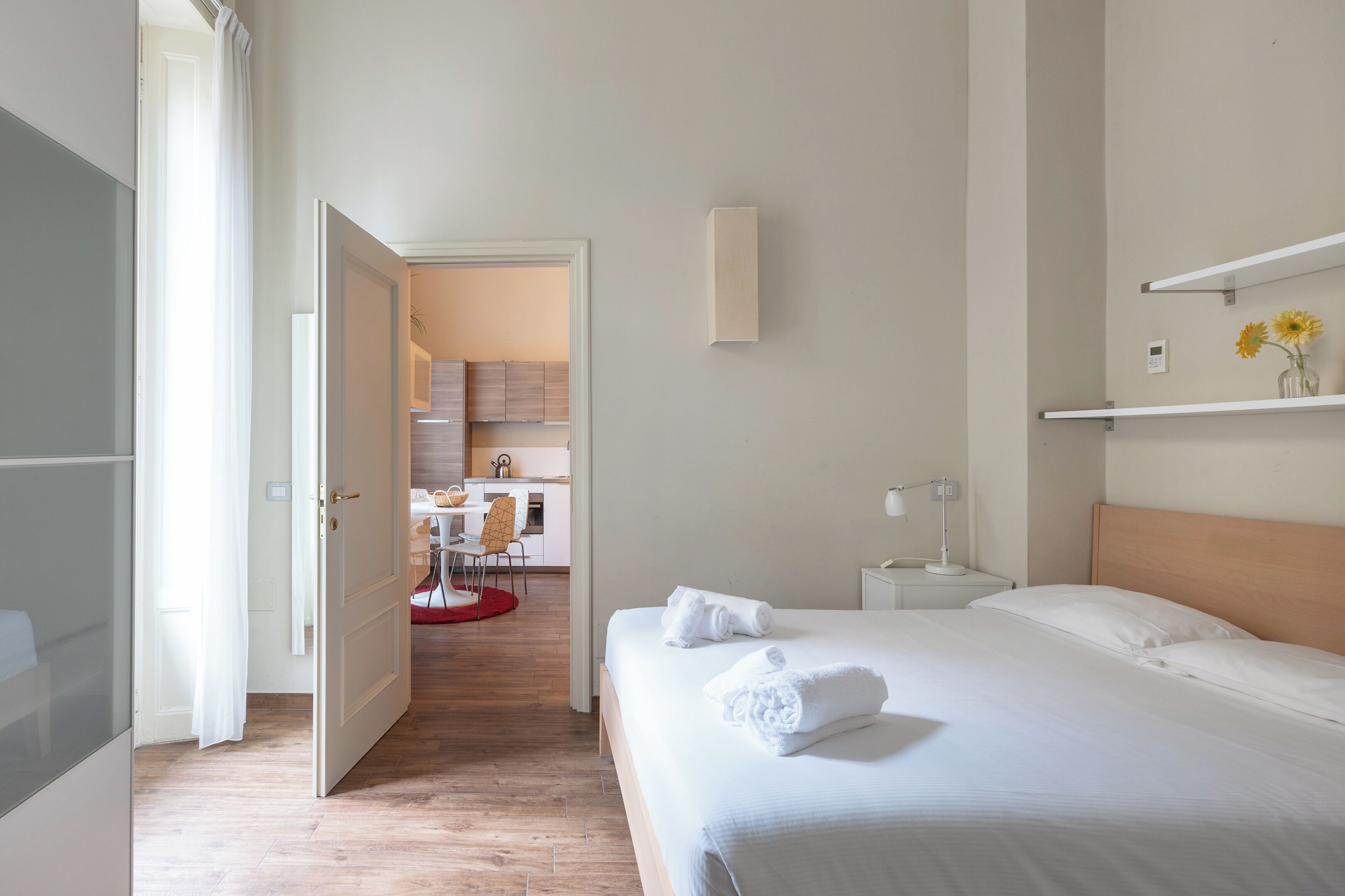 Quarto de hotel com cama, mesa de cabeceira, prateleira na parede com flores e vista para a cozinha.