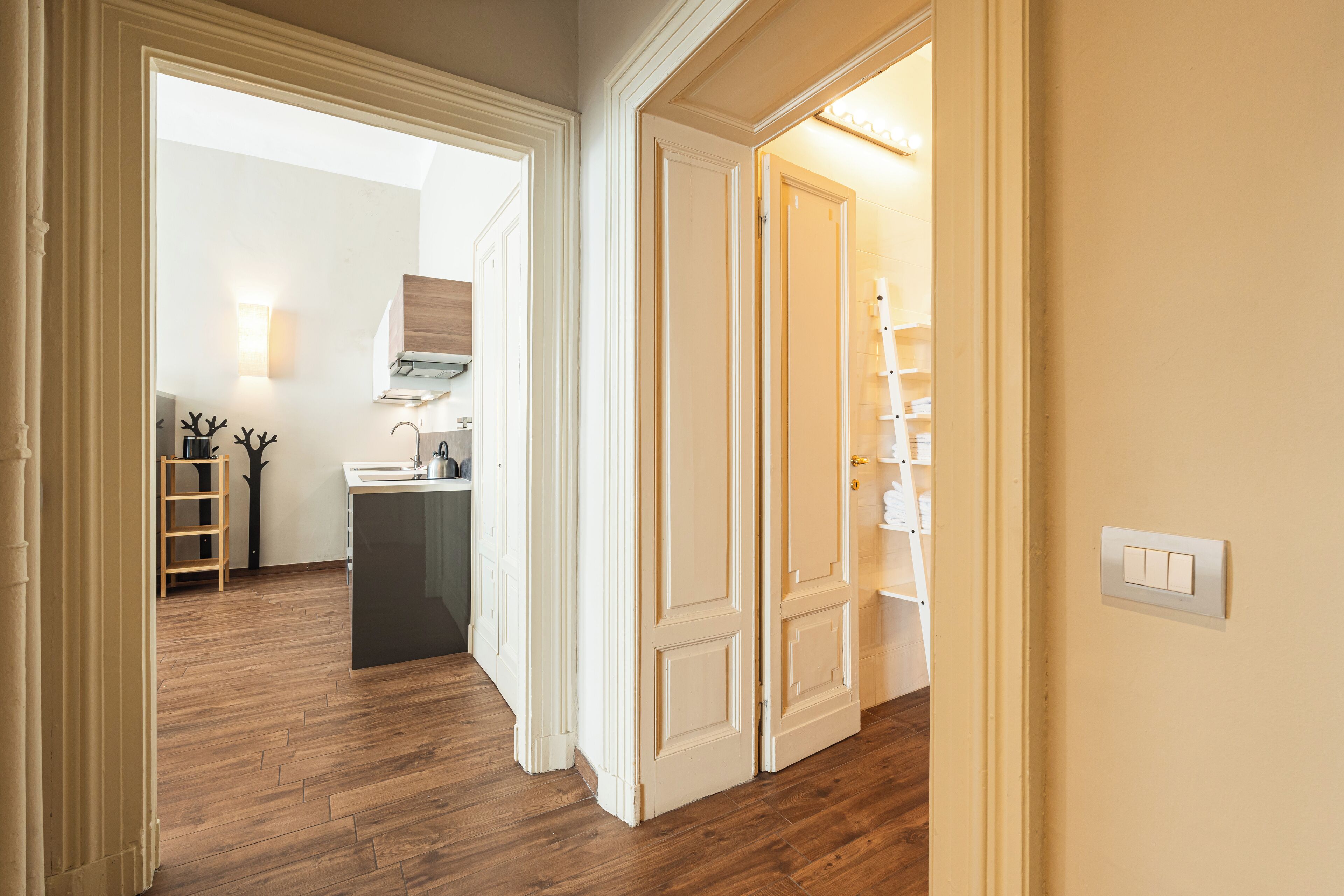 A hallway with wooden flooring, white walls, and a door leading to a kitchen area with a sink and cabinets.
