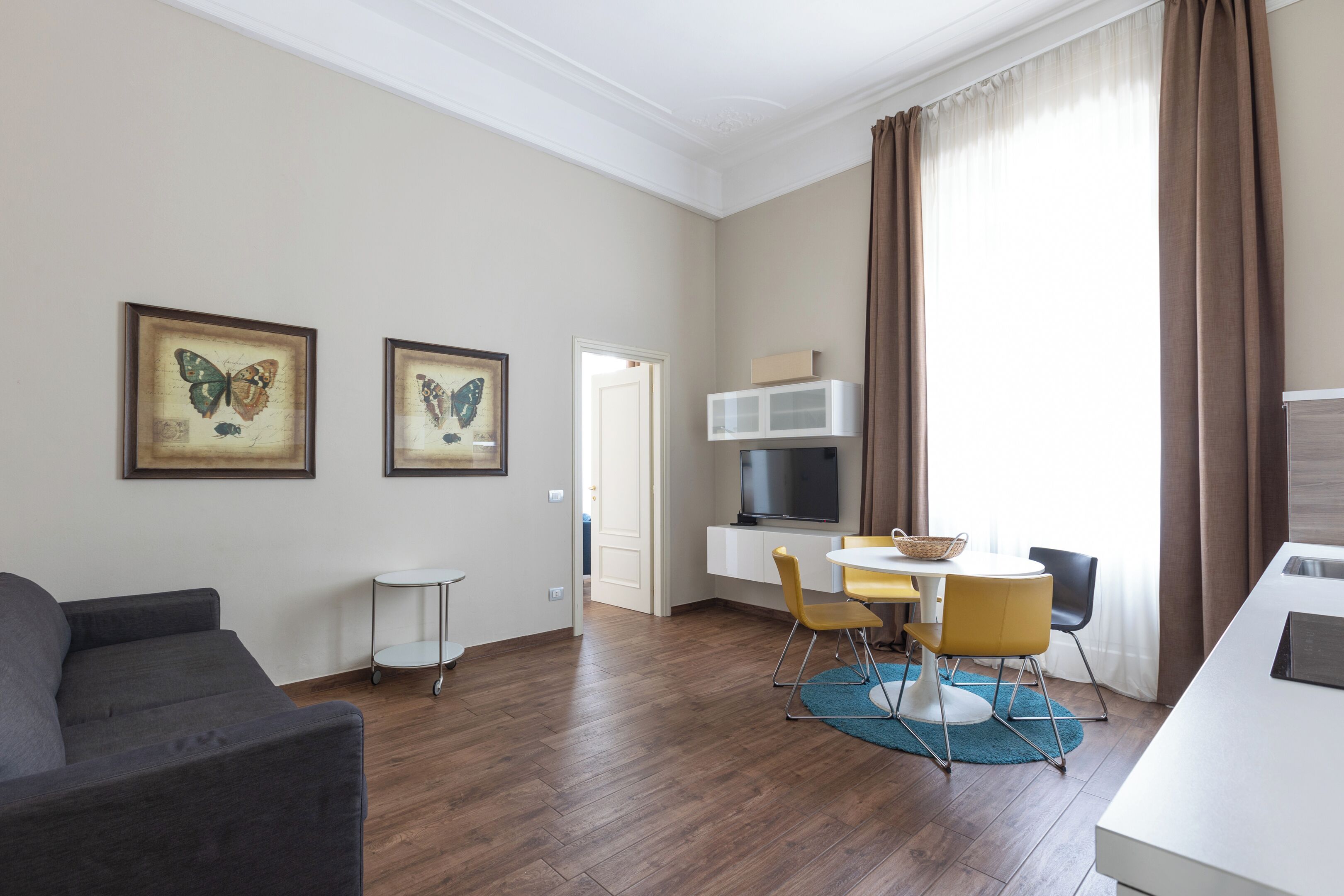 Una sala moderna con un sofá gris, una zona de comedor con sillas amarillas, un televisor sobre un mueble blanco y dos cuadros con mariposas en la pared.