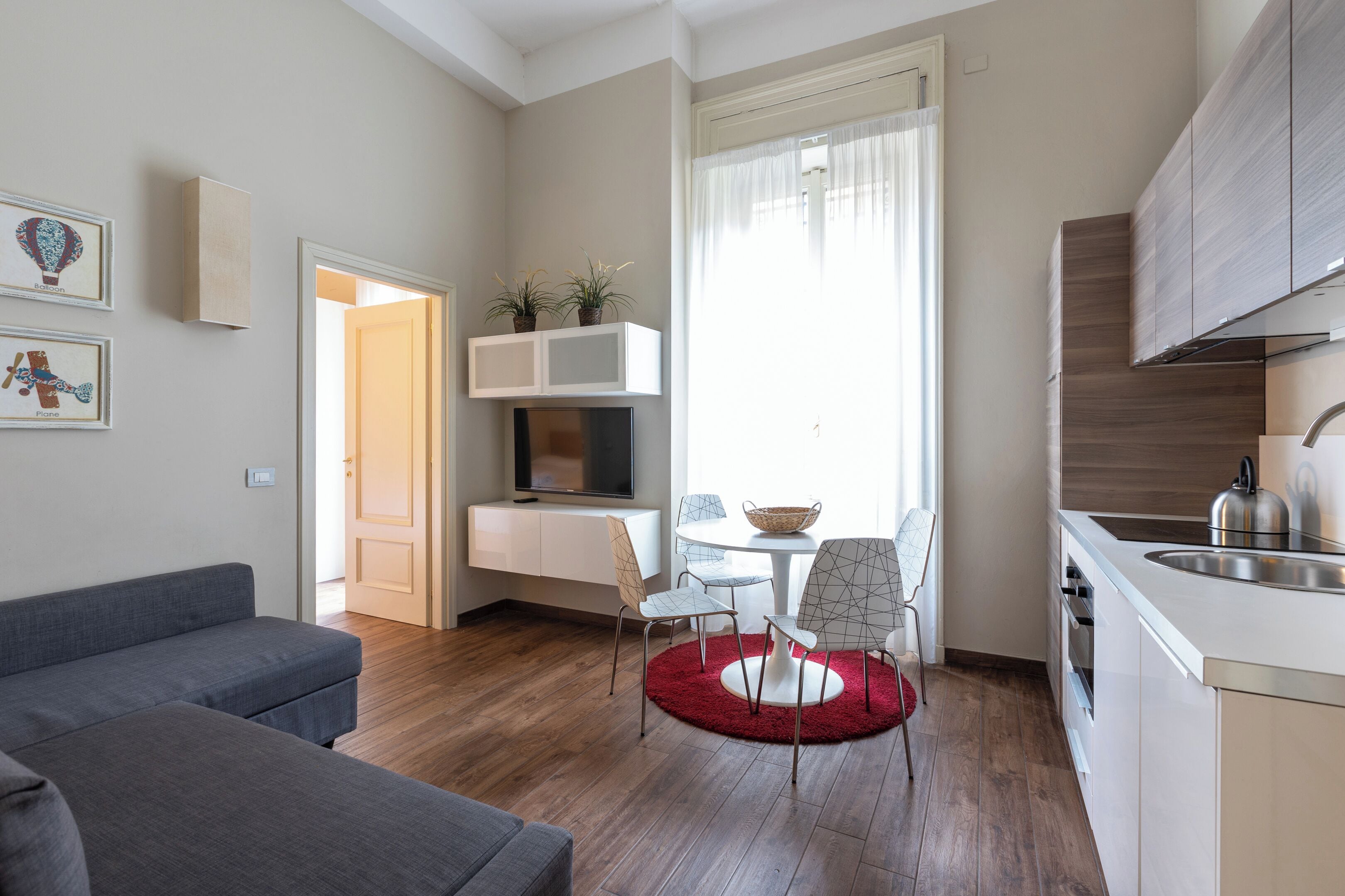 A modern living room with a grey sofa, a dining area with a red rug, and a kitchen with wooden cabinets and a stainless steel sink.