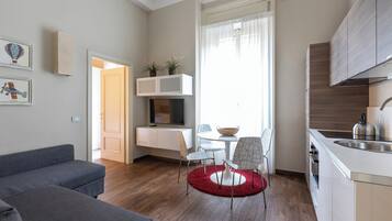 A modern living room with a grey sofa, a dining area with a red rug, and a kitchen with wooden cabinets and a stainless steel sink.