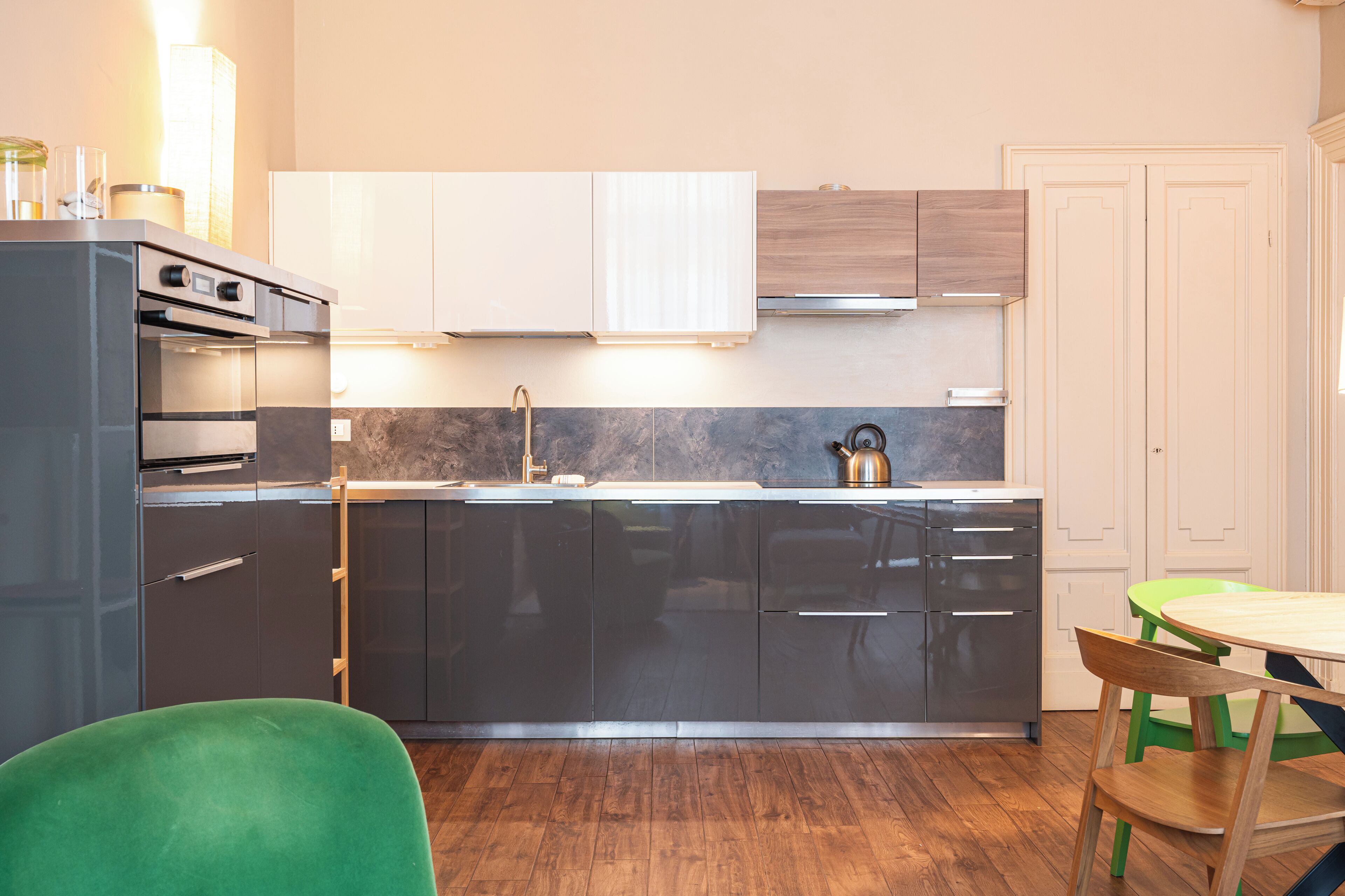 Una cocina moderna con horno empotrado, microondas y electrodomésticos de acero inoxidable. Hay una mesa de comedor de madera con dos sillas y un sillón de color verde.