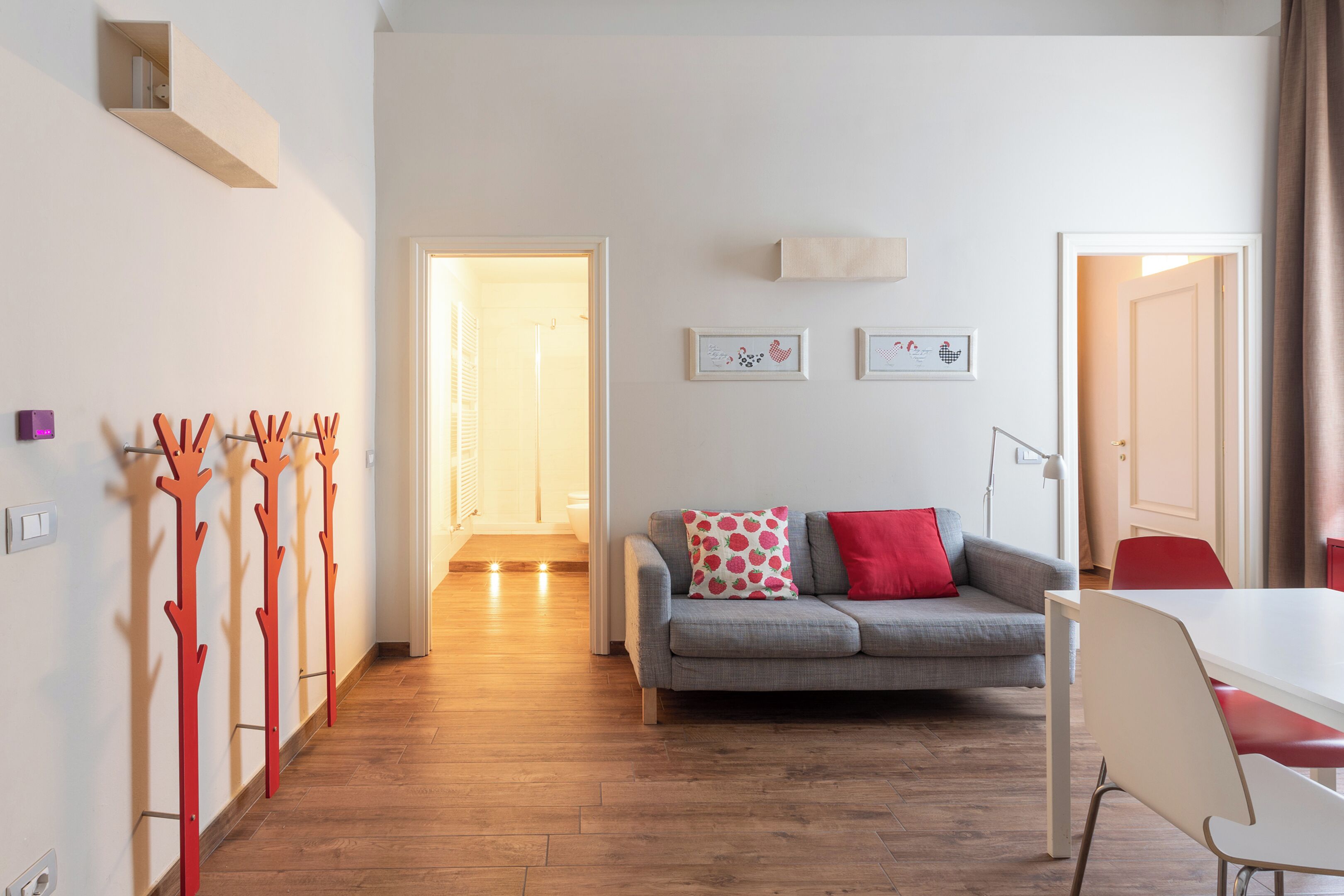A modern living room with a grey sofa, red and white pillows, a dining table with chairs, and a wooden floor.