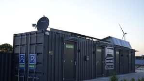 Un bâtiment modulaire avec des portes portant les inscriptions « ROATEL » et « SLEEP WELL », une antenne parabolique et une éolienne en arrière-plan.