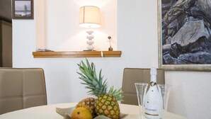 Ruokapöydällä on hedelmäkulho, jossa on ananas, appelsiini ja banaani. Pöydällä on myös pullo Chenet-viiniä ja useita lasia. Pöydän yläpuolella olevalla hyllyllä on lamppu, jonka varjostin on beigen värinen.