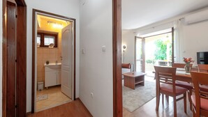 A modern kitchen with a sink and cabinets, a dining area with wooden furniture, and a view of the outdoors through a large window. - Madonna Field Apartment II - Peaceful 1BR for 3 in Lapad (Dubrovnik)