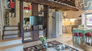 Ein Wohnzimmer mit Holzboden, einem Fernseher, einem Essbereich mit Tisch und Stühlen sowie einer Küche im Hintergrund.