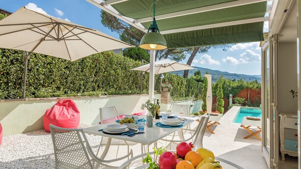 Eine Terrasse im Freien mit Esstisch, umgeben von weißen Stühlen und Sonnenschirmen, mit Blick auf einen Pool und eine Berglandschaft.