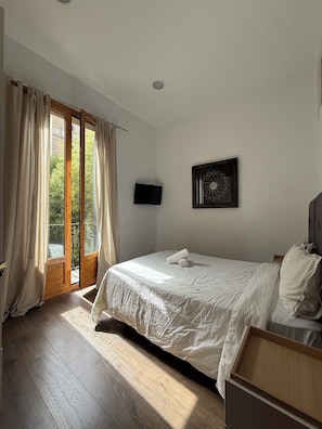 A bedroom with a large bed, a flat-screen TV, a window with curtains, and a view of greenery outside. - Bright Central Apartment (Barcelona)