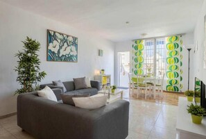 A modern living room with a sofa, a painting, a potted plant, and a dining area with a patterned curtain.