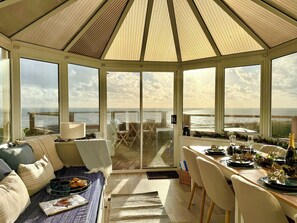A sunlit room with a view of the sea, a dining area with a table set for two, and a cozy seating area with cushions and a tray of pastries. - Crows Nest Dawlish (Dawlish)