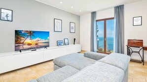 Een moderne woonkamer met een flatscreen televisie, grijze banken, een witte tv-kast en uitzicht op zee vanaf een balkon.