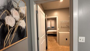 A hallway leading to a bedroom with a bed, a nightstand, and a window with blinds. There is a painting on the wall and a light switch on the wall.