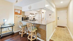 A compact kitchen with white cabinets, a microwave, and a sink. There are three wicker bar stools with green cushions. A small desk with a lamp is visible to the left. - Heron Pointe D5- 2BR- 5 Minute Walk To Beach (Myrtle Beach)