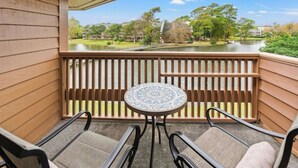 A balcony with a view of a lake, wooden railing, and a small round table with two chairs. - Kingston Laurel Court 311- 2BR/2BA- Outdoor Pool (Myrtle Beach)