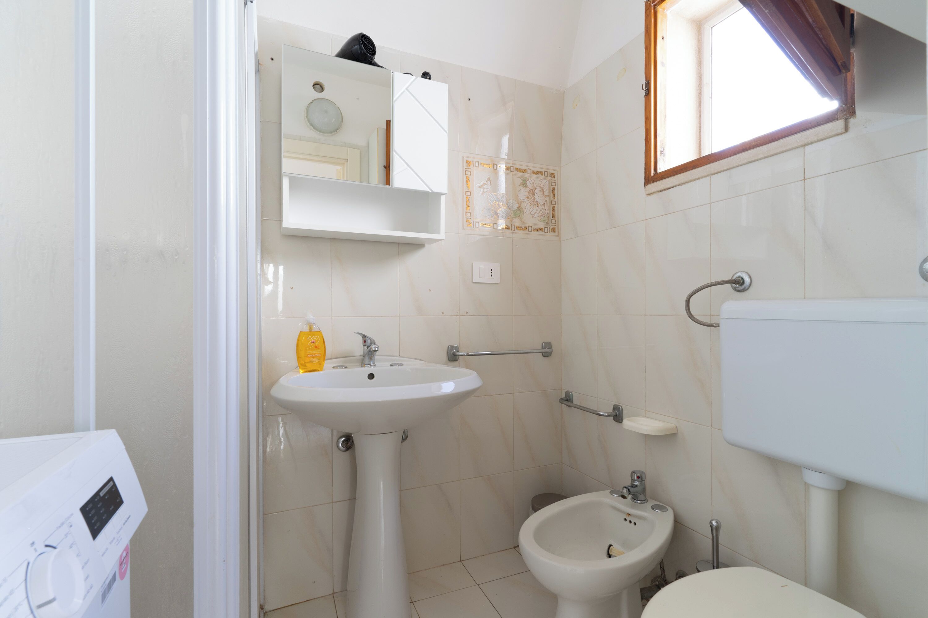 A small bathroom with a pedestal sink, a bidet, a mirror, and a window.
