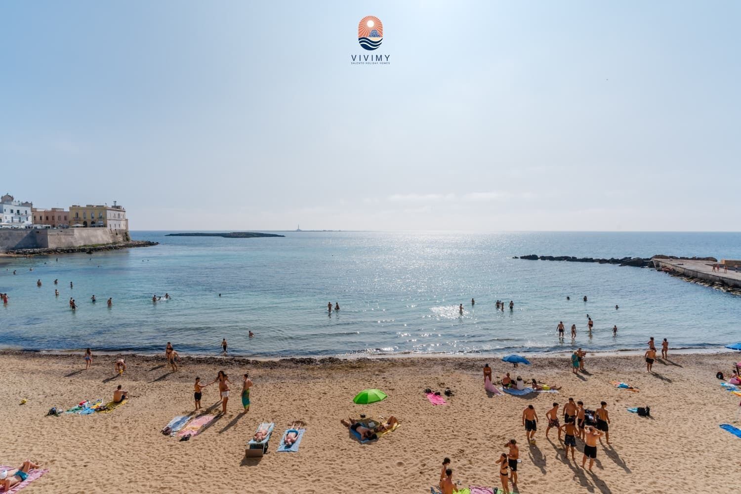 Una playa con gente nadando y tomando el sol, un muelle y edificios al fondo.