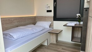 Un dormitorio pequeño con una cama individual, una mesita con toallas, una planta en maceta y una ventana con persianas.