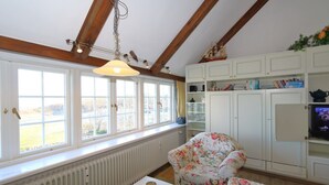 Een kamer met een groot raam, houten balken, een bloemen fauteuil, een radiator en een televisie.
