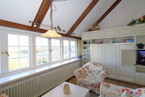 Ein Zimmer mit großem Fenster, Holzbalken, einem blumigen Sessel, einem Heizkörper und einem Fernseher.
