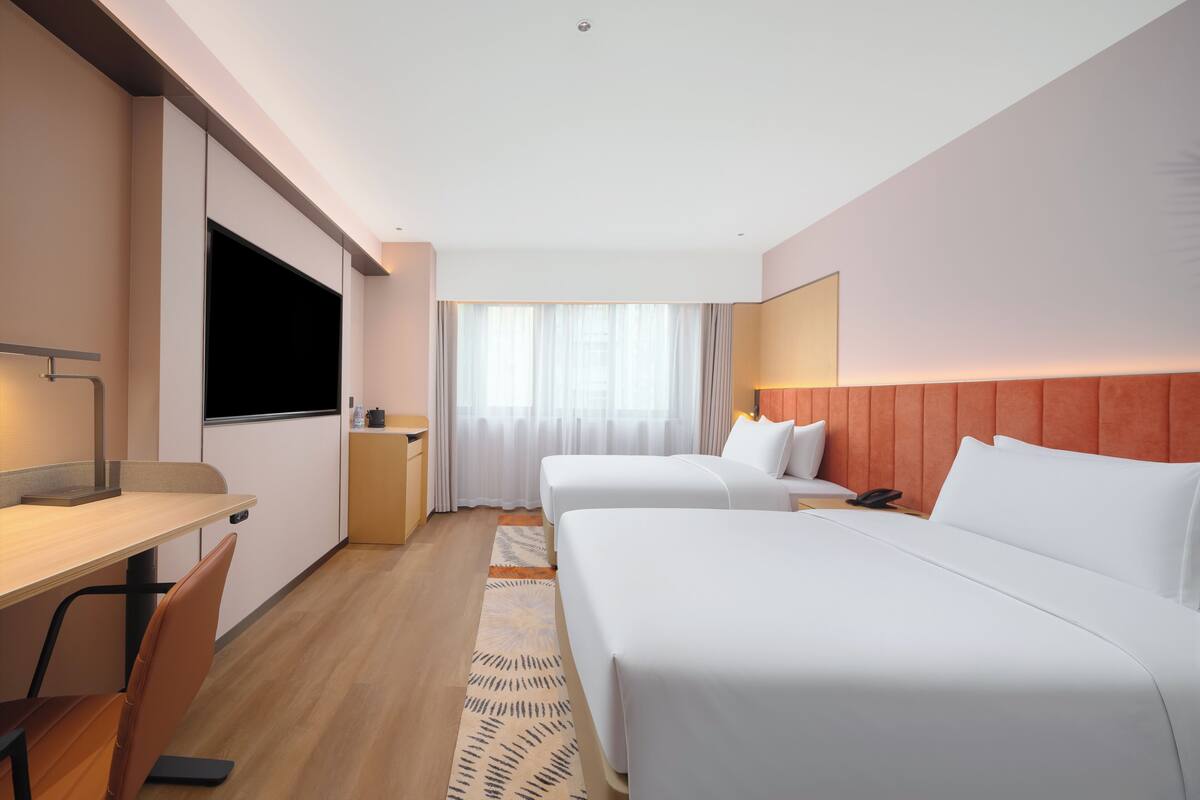 A hotel room with two beds, a desk, a TV, and a window with curtains.