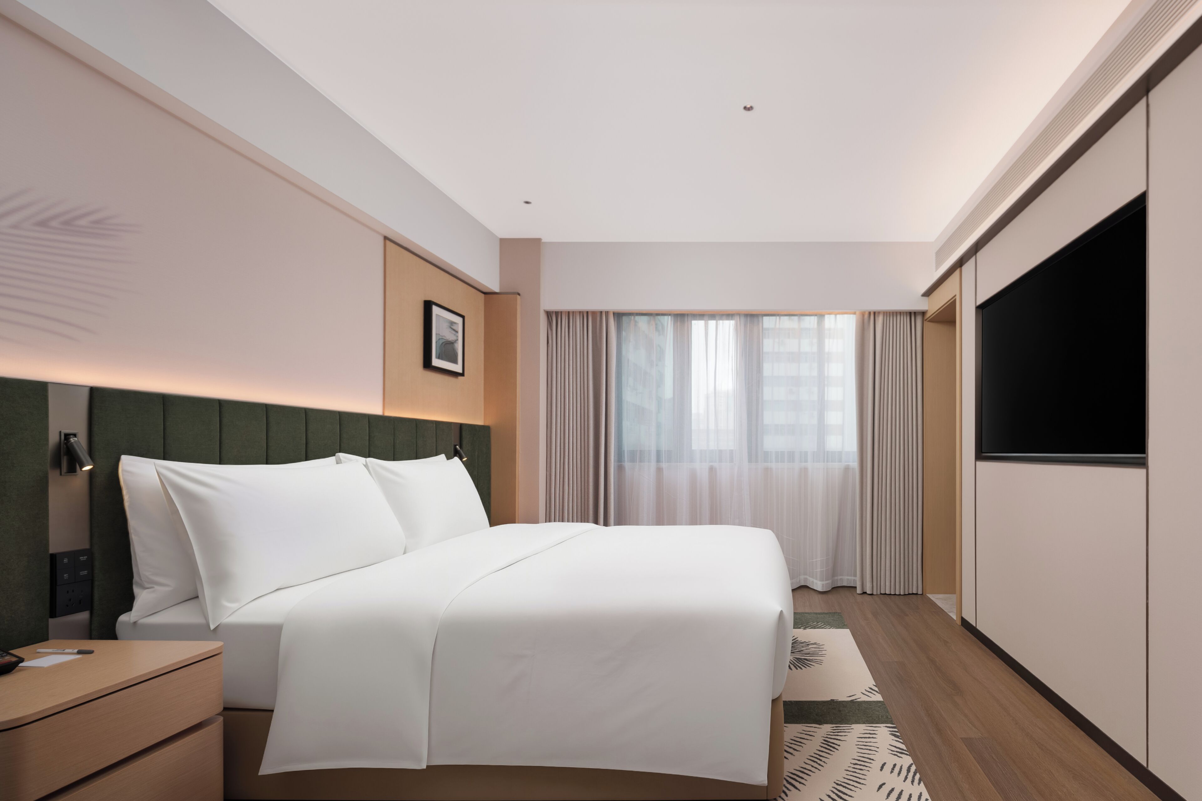 Une chambre d’hôtel équipée d’un lit, d’une télévision, d’une table de chevet, d’une fenêtre avec des rideaux et d’une œuvre d’art accrochée au mur.