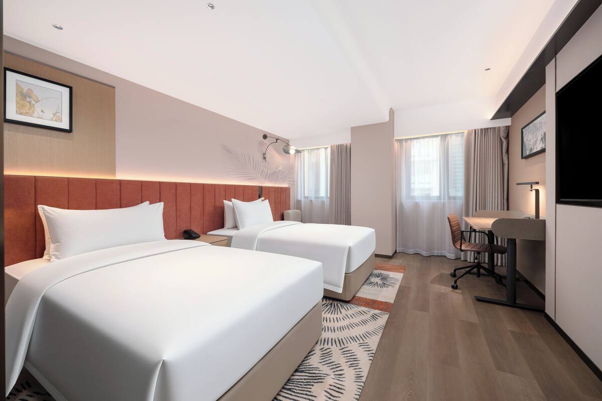 A hotel room with two beds, a desk, a chair, a TV, and a window with curtains.