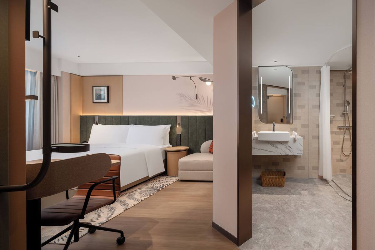 A hotel room with a bed, desk, chair, bedside table, lamp, and a bathroom with a shower and sink.