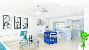 Ein modernes Wohnzimmer mit einem weißen Sofa, einem Glastisch und einem blauen Akzentstuhl. Es gibt eine Küchenzeile mit einer weißen Kochinsel und einem großen Fenster.