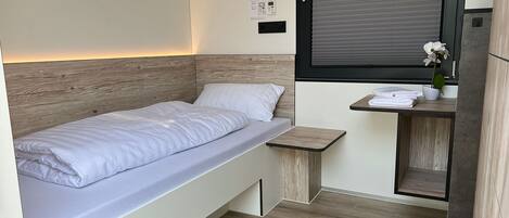Quarto compacto com uma cama de solteiro, uma mesinha com toalhas, uma planta em vaso e uma janela com persianas.