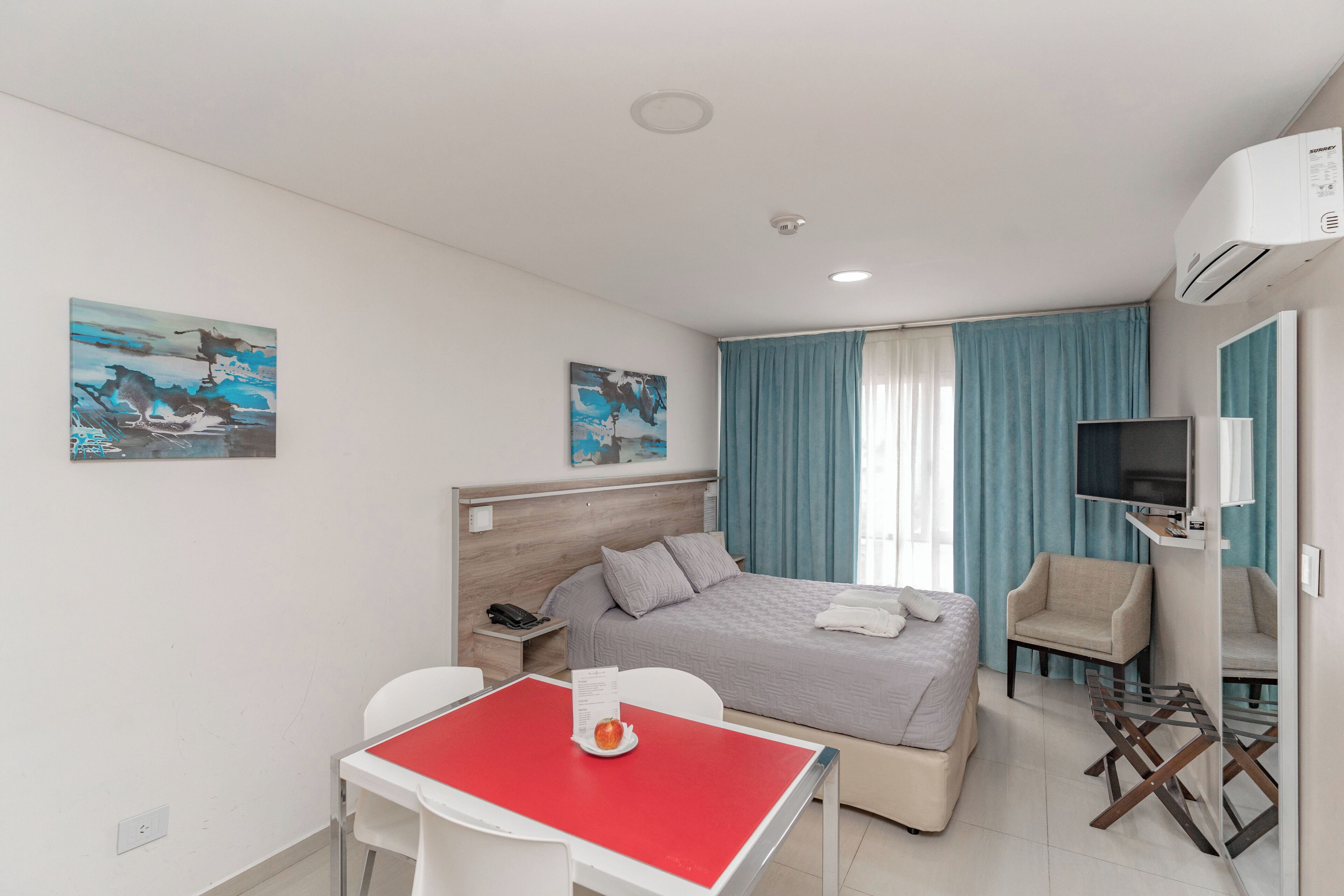 A hotel room with a bed, a TV, a desk with a red table, a chair, a sofa, and artwork on the walls.