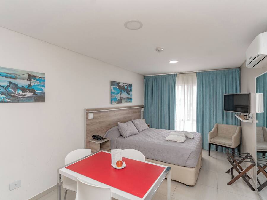 Et hotelværelse med seng, fjernsyn, skrivebord med rødt bord, stol, sofa og kunst på væggene.