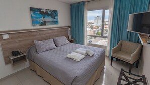 Une chambre d’hôtel avec un grand lit, une chaise, une télévision, un tableau et une fenêtre avec des rideaux.
