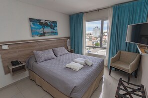 Une chambre d’hôtel avec un grand lit, une chaise, une télévision, un tableau et une fenêtre avec des rideaux.