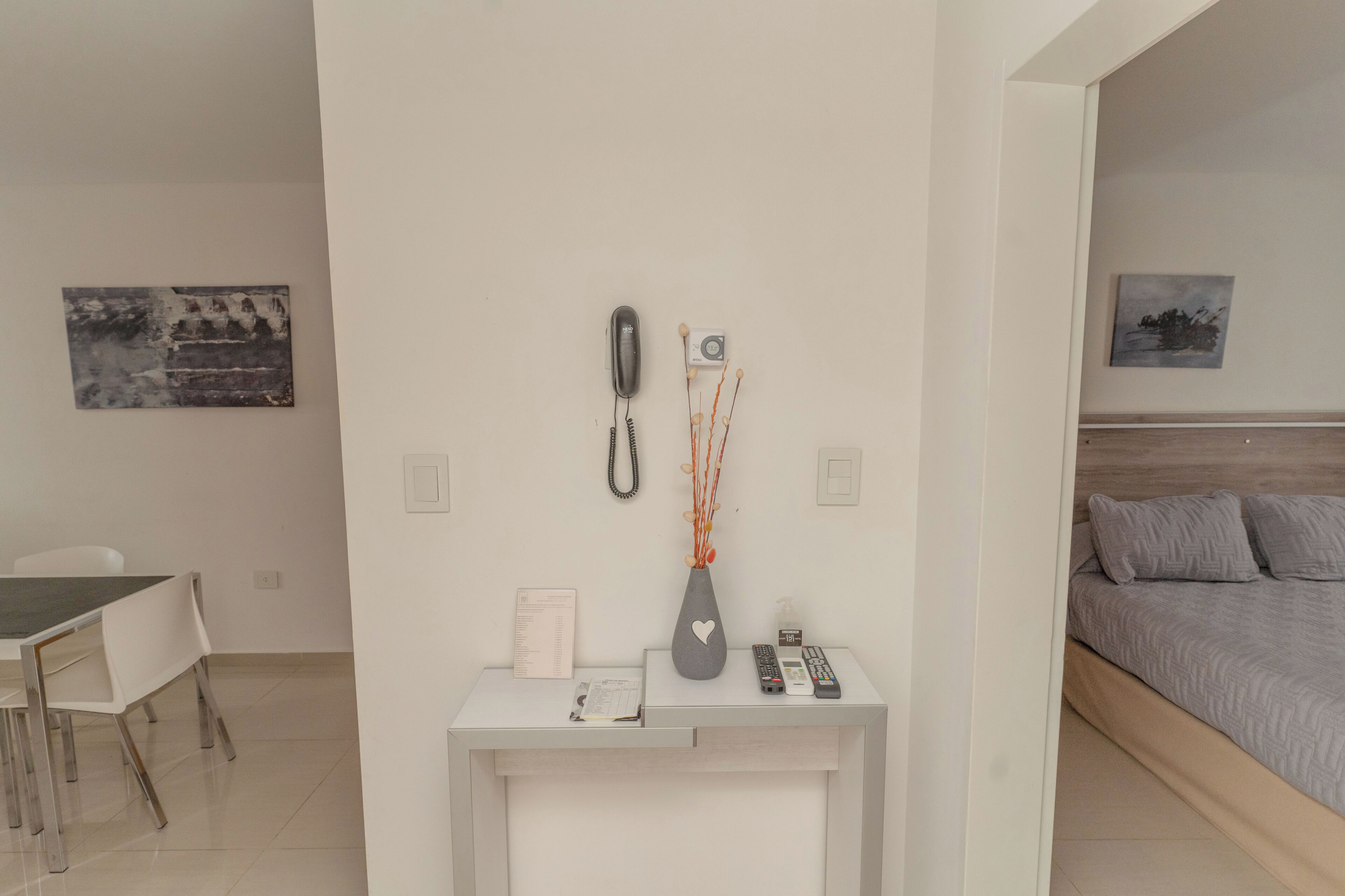 A modern hotel room with a white desk, a bed with grey bedding, a small vase with branches, and a phone on the wall.