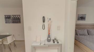 A modern hotel room with a white desk, a bed with grey bedding, a small vase with branches, and a phone on the wall.