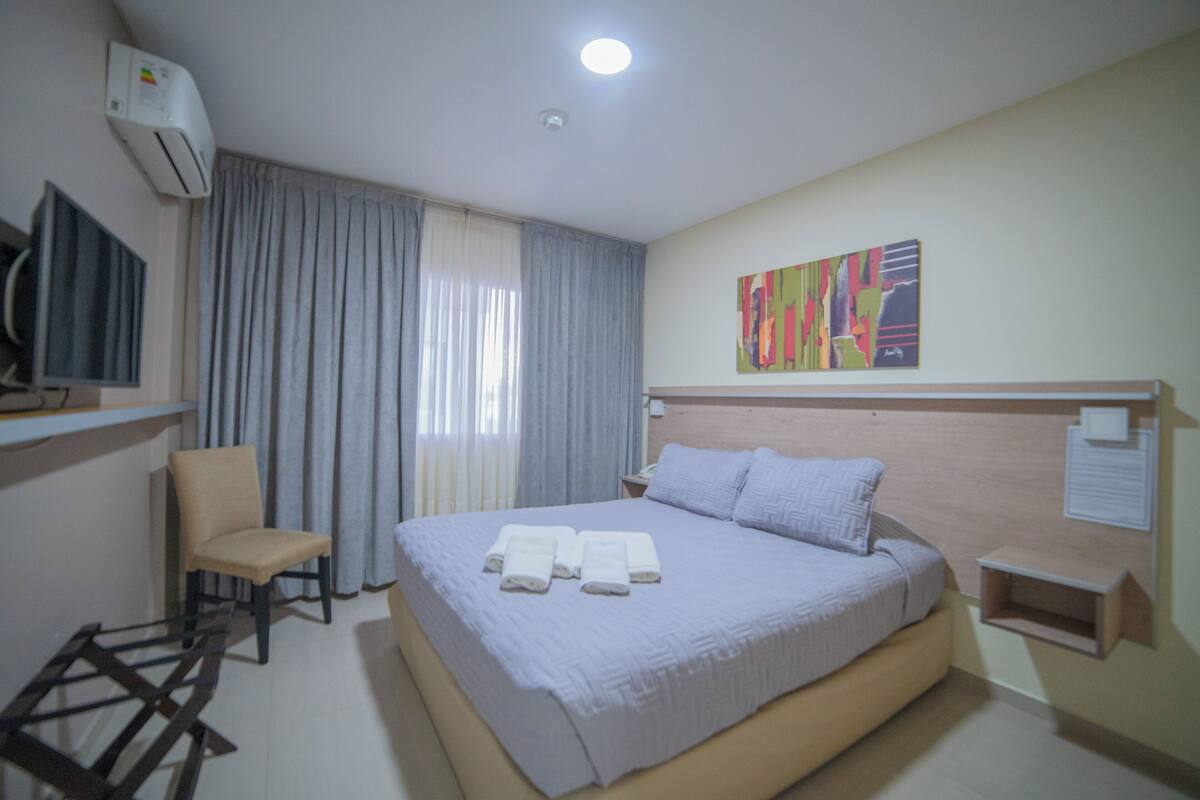 Een hotelkamer met een bed, een stoel, een airconditioner, een schilderij en een tafeltje.