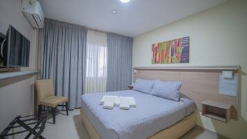 Una habitación de hotel con cama, una silla, aire acondicionado, un cuadro y una mesita.