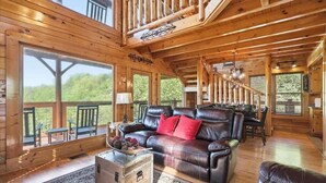 Interior de uma cabana de madeira com sofá de couro, área de refeições e janelas amplas.