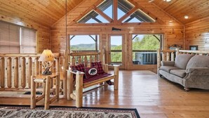 A log cabin interior with a wooden ceiling, a sofa, a bench, and a view of the outdoors through large windows.