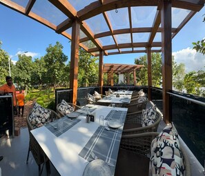 An outdoor dining area with wooden pergola, tables set with white placemats, and wicker chairs with patterned cushions. - ODHNI DAM RESORT (Banka)