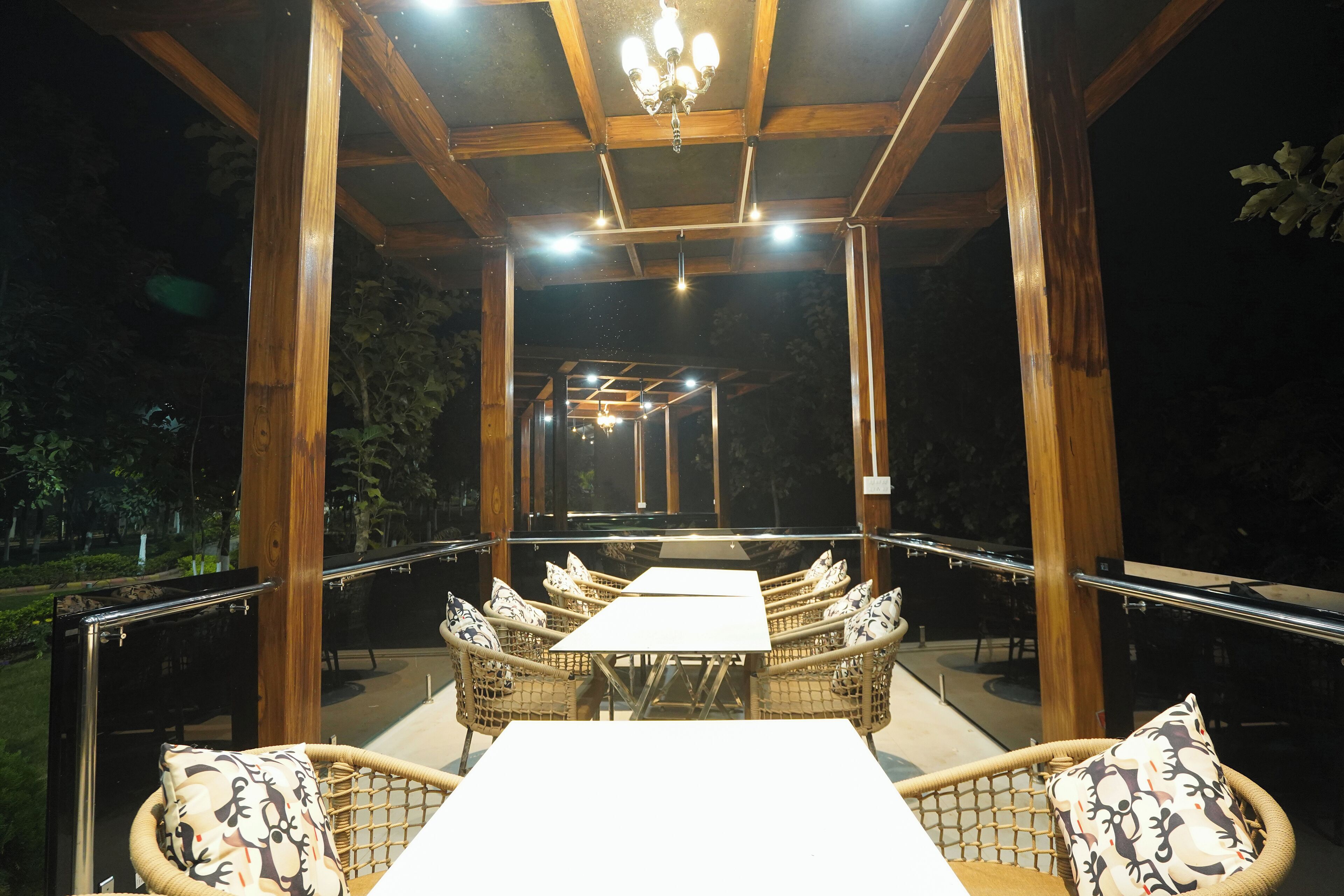 Un patio techado al aire libre con una mesa larga y sillas de mimbre, rodeado de vigas de madera y una barandilla de vidrio.